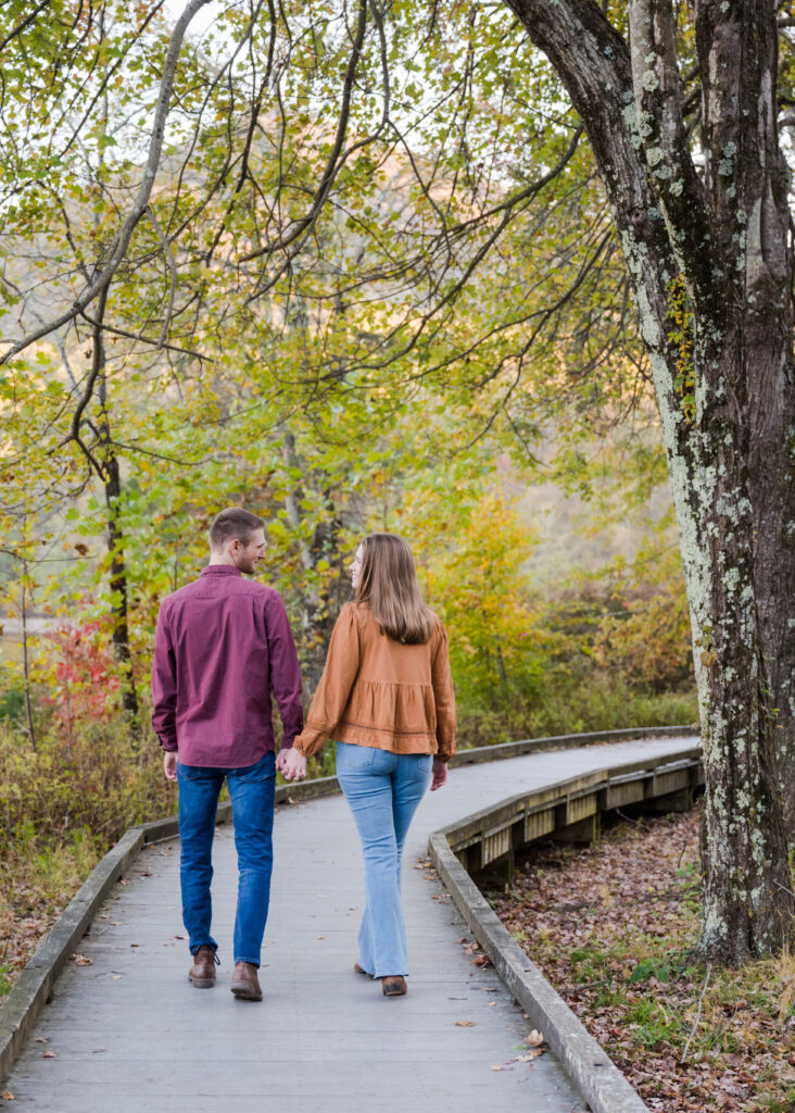 A couple walking hand in hand down a tree-lined wooden pathway during their engagement session. The man, wearing a burgundy shirt and jeans, and the woman, in an orange blouse and light blue jeans, stroll peacefully under the canopy of green leaves. The vibrant autumn foliage creates a colorful, serene backdrop, with shades of yellow, orange, and red peeking through the trees. Their backs are turned to the camera, adding a sense of togetherness and quiet connection as they enjoy this peaceful moment in nature.