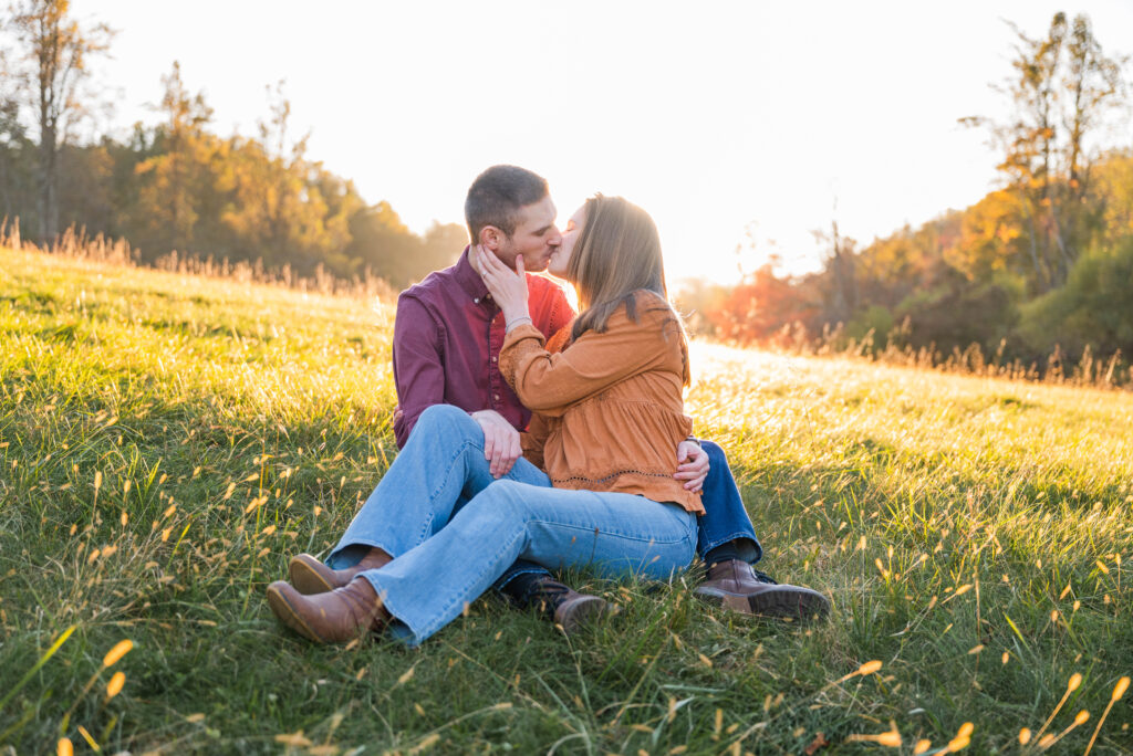 A romantic moment captured during an outdoor engagement session at sunset. The couple sits closely together in a grassy field, sharing a tender kiss. The man, dressed in a burgundy shirt, wraps his arms around the woman, who is wearing an earthy orange blouse and light jeans. The golden sunlight bathes the scene, casting a warm glow over the couple and the surrounding meadow, with trees in the background softly illuminated by the fading light. The image exudes love, warmth, and serenity.