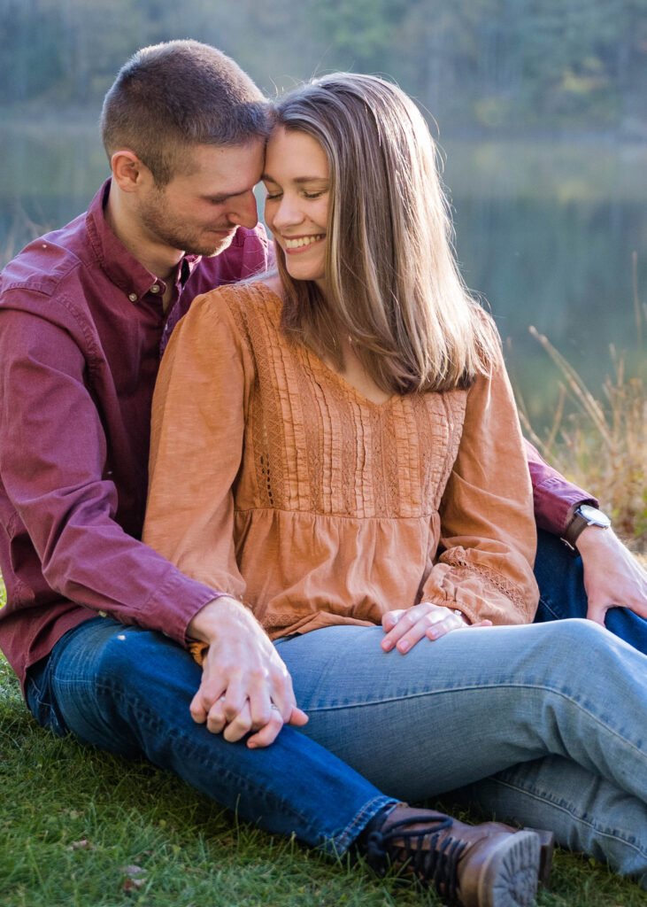 
A sweet and intimate moment between a couple during their engagement session. The man, wearing a burgundy button-up shirt, sits closely behind the woman, who is dressed in an earthy orange blouse and light blue jeans. They are seated on the grass, holding hands, and smiling as they lean their heads together. The warm sunlight softly illuminates them, with a serene lake and forested background adding to the peaceful, romantic atmosphere.